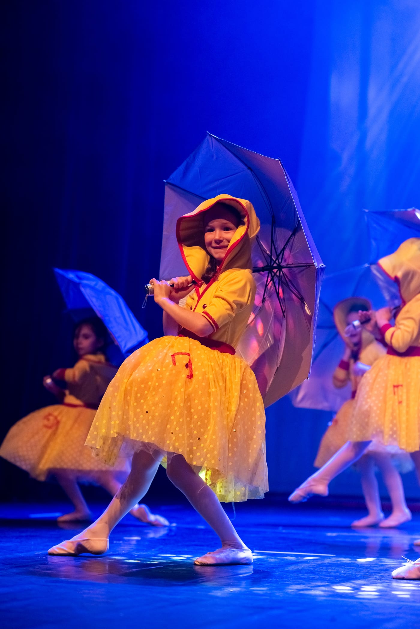 Menina de figurino amarelo sorrindo no palco, segurando um guarda-chuva